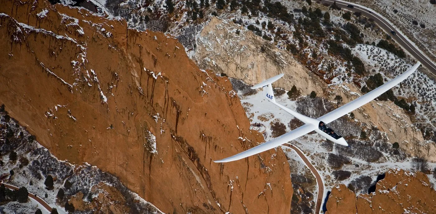 A white sailplane banking hard over red-ochre ridges, wings stretched across the whole sky