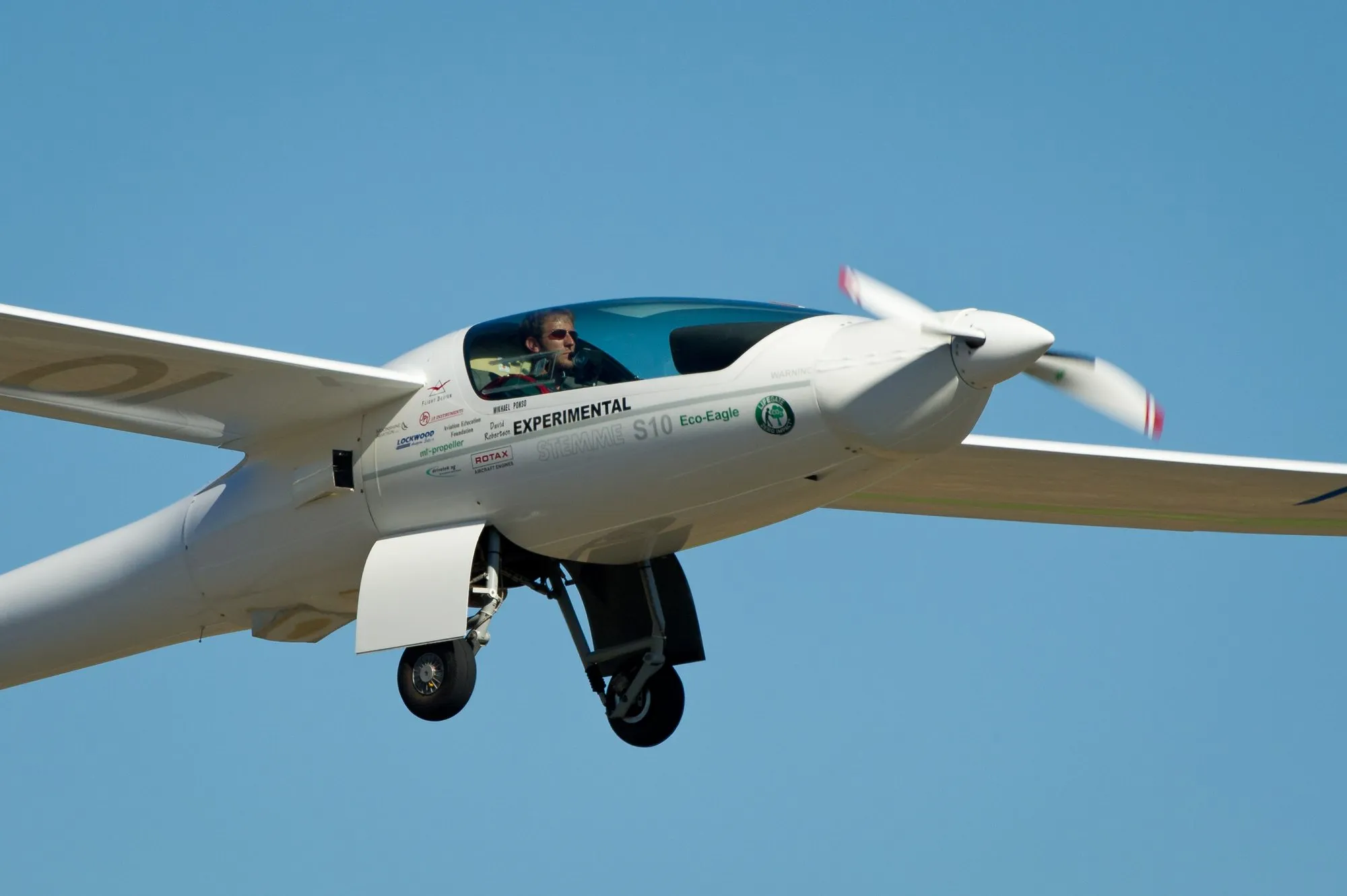 A Stemme S10 motor glider just after take-off, propeller blurred, pilot visible under the long canopy